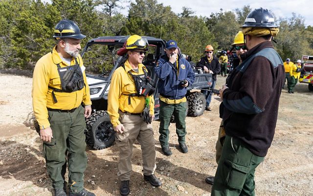 A group of men dressed in yellow fire gear and helmets stand in front of an ATV talking.